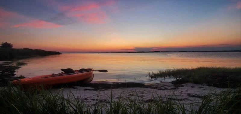 A kayak resting on a sandy beach at sunset with a calm lake and a colorful sky in the background.