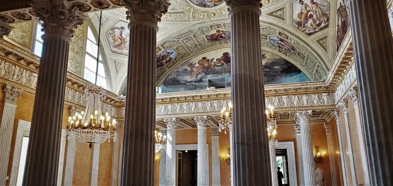 Elegant interior with large columns, ornate ceiling murals, chandeliers, and decorative molding.