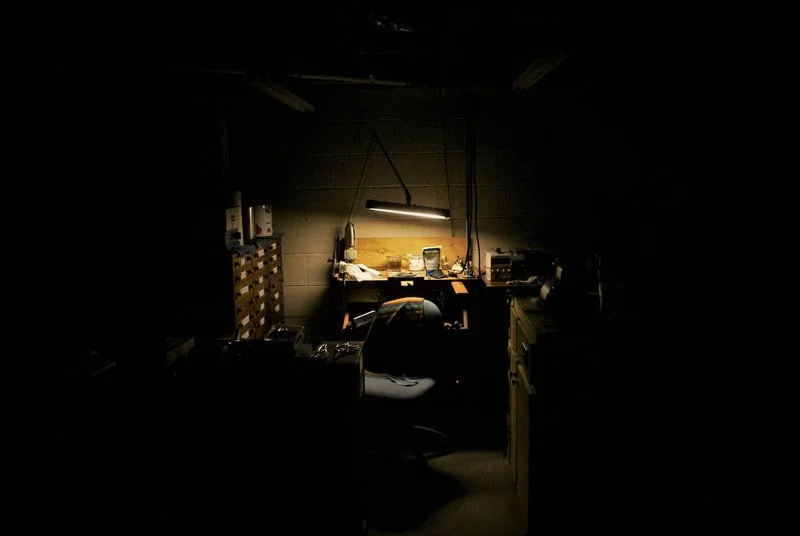 Dimly lit workspace with a desk, a chair, and a fluorescent light above, surrounded by various tools and equipment.