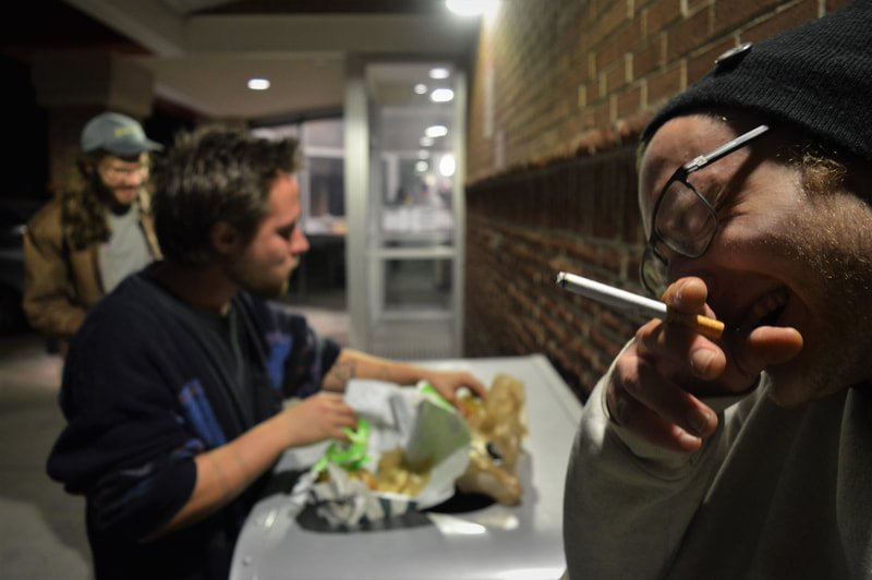Three young men at a fast food counter, one smoking a cigarette, with a brick wall in the background.