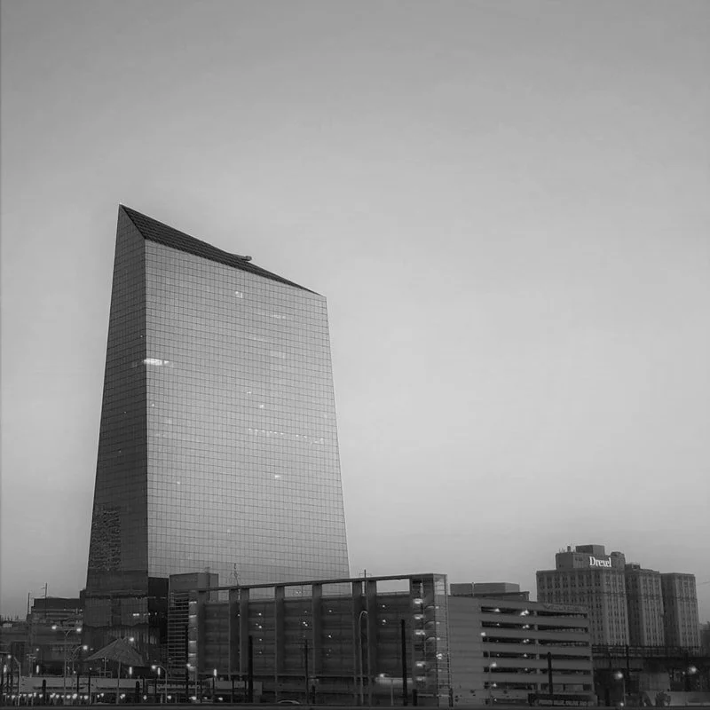 A tall modern glass skyscraper with a curved top in an urban cityscape, with other buildings and a parking structure in the foreground, in black and white.