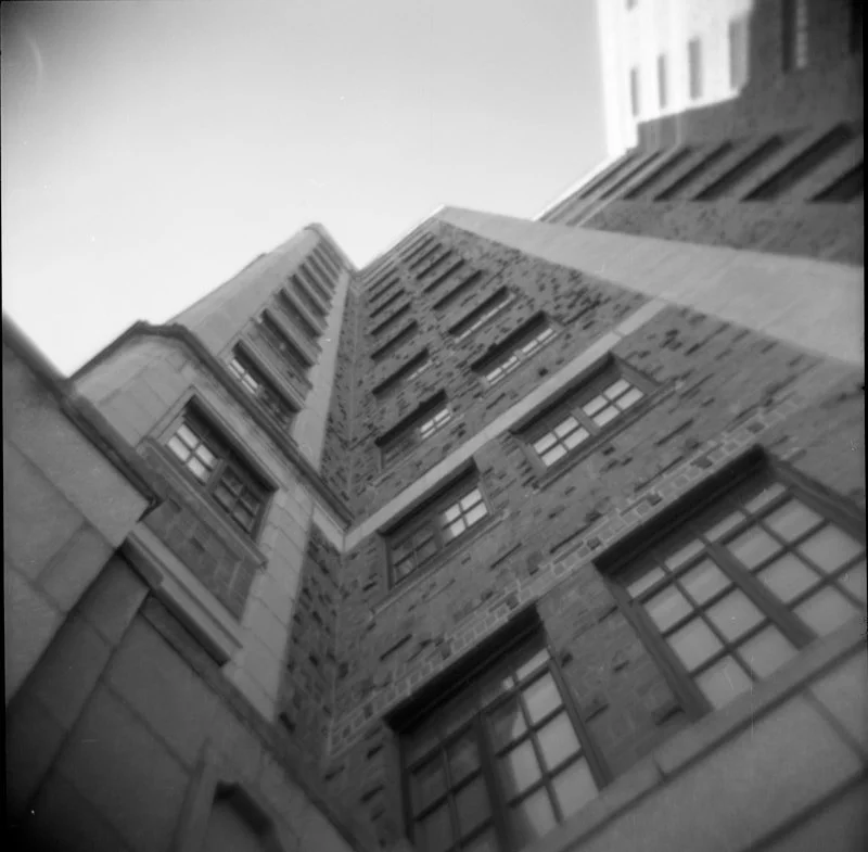 Black and white photo of a tall, multi-story brick building taken from the ground looking up, with a clear sky in the background.