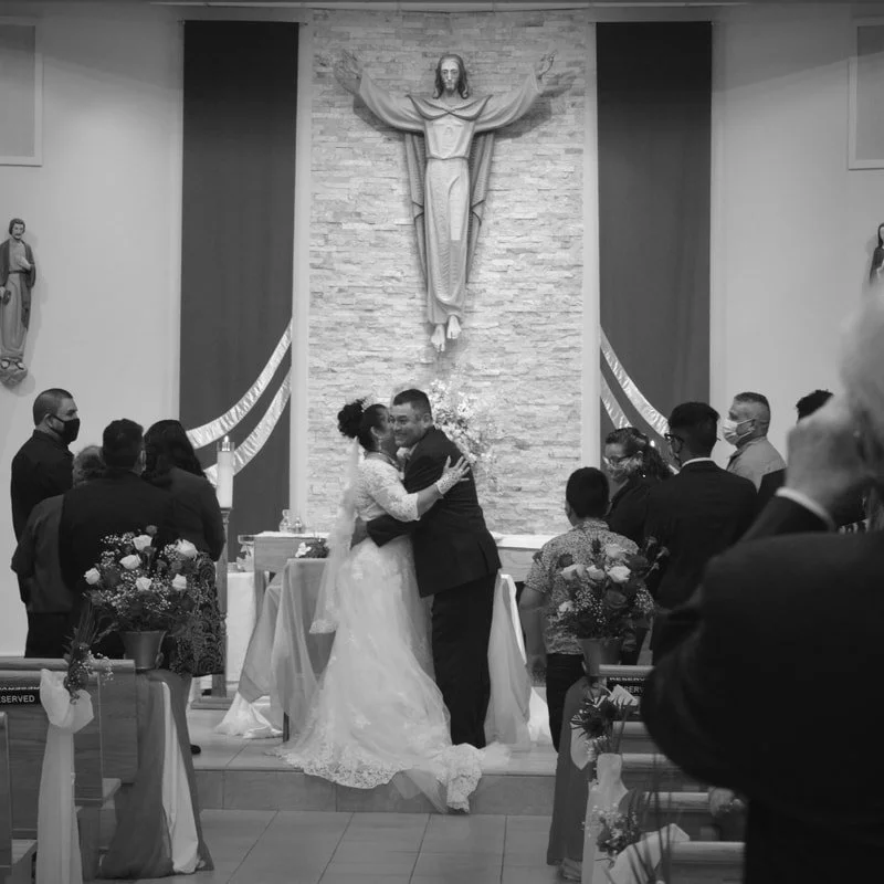 A wedding ceremony with a bride and groom hugging at the altar in a church, with religious decorations and guests in the background.