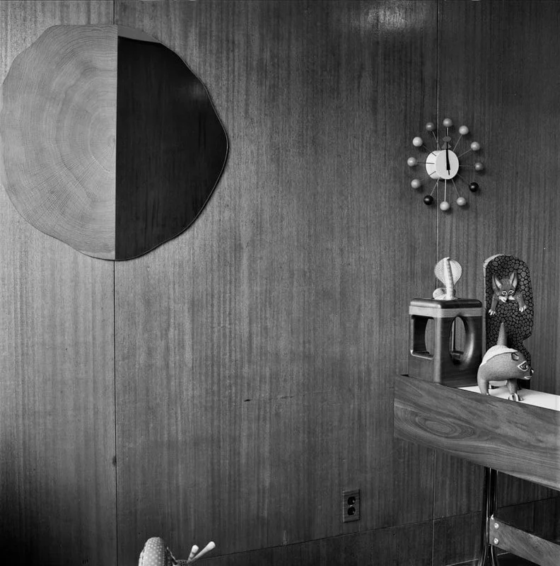 A wooden wall with a round abstract wall art on the left, and a vintage clock with a sphere design on the right. Below the clock, a wooden shelf holds various decorative figurines, including a woman with long hair, a cat, and a pig.