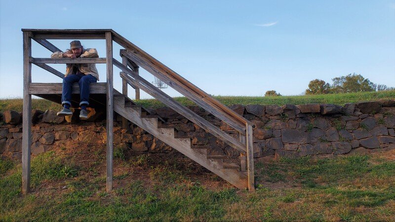 A person sitting on a wooden observation platform at the top of stairs in a grassy area with a stone wall and trees in the background.
