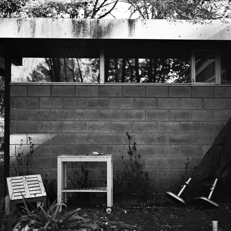 Black and white photo of a backyard scene with a concrete block wall, a small wooden table, garden plants, a stack of wooden slats, and two stacked chairs partially covered with a tarp.