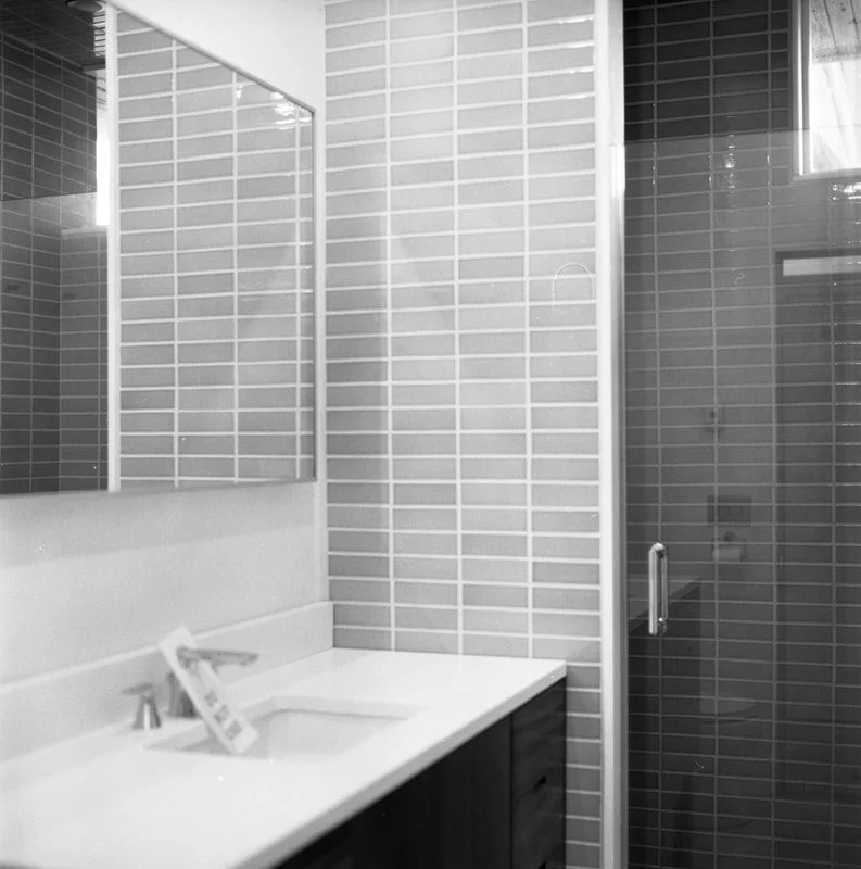 A modern bathroom with a rectangular mirror, light-colored tiled walls, a white countertop with an integrated sink, and a glass shower door with a metal handle.
