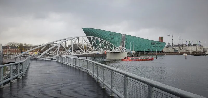 A modern building with a unique green exterior and a white bridge over a body of water.