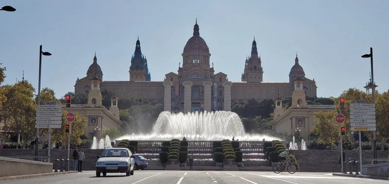 View of a grand historic building with domes and spires, with a fountain and steps in the foreground, and cars and people in the street.