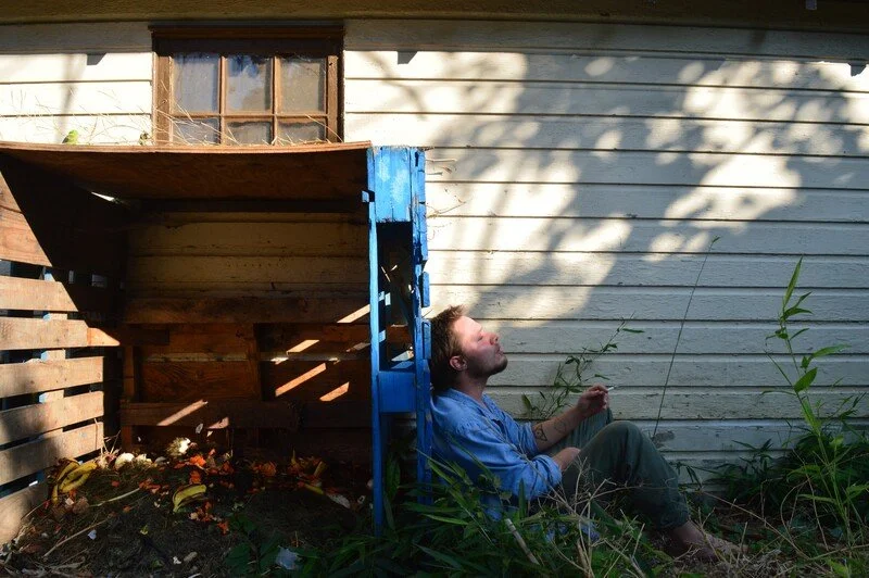 A man in a blue shirt sitting on the ground leaning against a house exterior, surrounded by grass and plants, with a wooden staircase and window behind him. Shadows of tree branches cast over the wall.