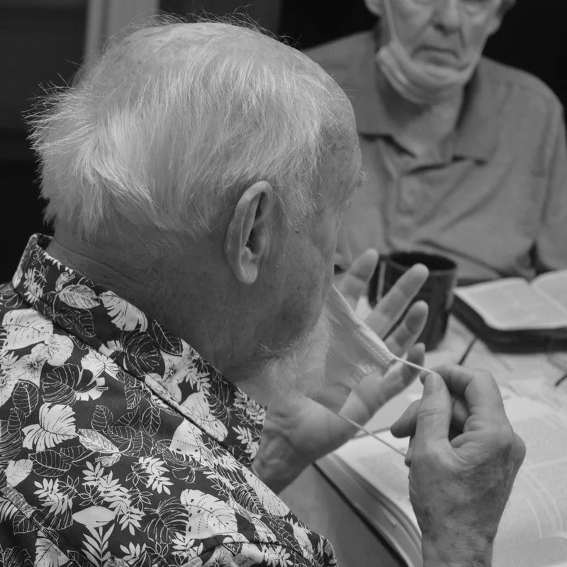 Two elderly men sitting at a table with books and cups, one is holding a glass with a straw and the other is wearing glasses and reading a book.