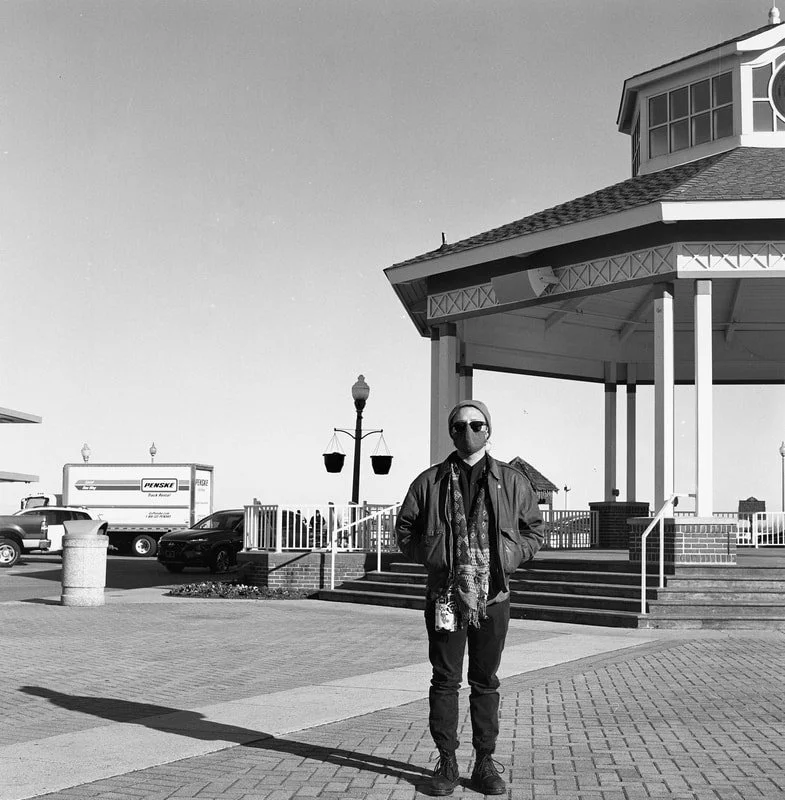 A man wearing sunglasses, a jacket, and a scarf standing outdoors near a gazebo with stairs, lampposts, and parked cars in the background.