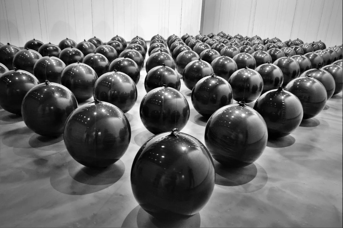 Black spherical objects arranged in rows on a smooth floor in a gallery or exhibition space.