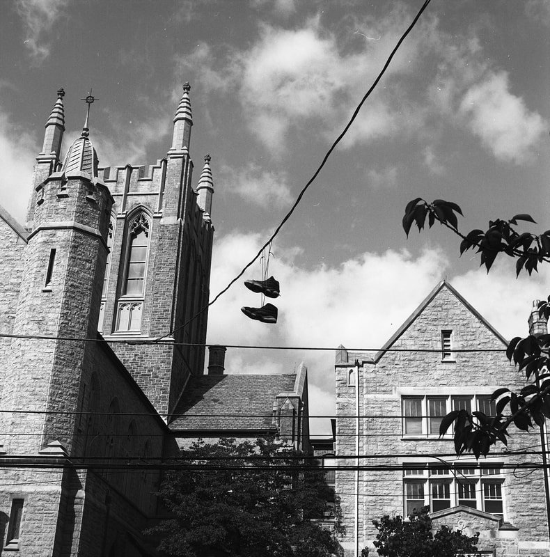 Black and white photo of a historic church or cathedral with Gothic architecture, tall towers, and large stained glass windows. Clothes are hanging to dry on a rope across the foreground, with some leaves visible on the right side of the image.