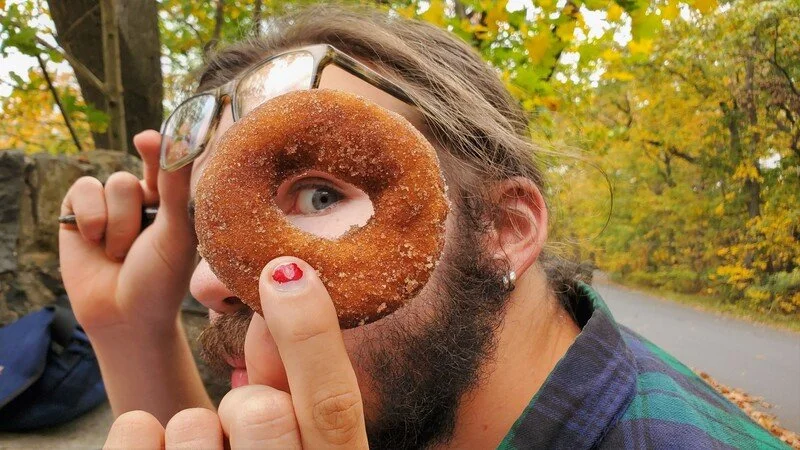 A man with glasses and a beard holding a glazed donut with a hole in front of his face, making it look like an eye, outdoors surrounded by autumn trees.