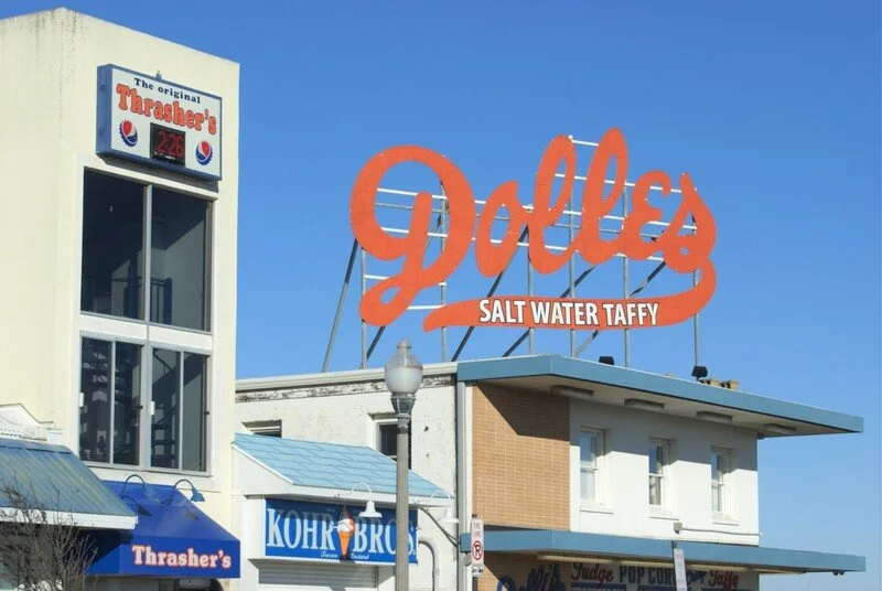 Sign for Dolores Salt Water Taffy store with large, orange lettering and a smaller blue and red sign advertising Thrasher's fries on the side.