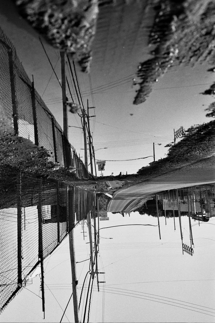 Black and white photo of a flooded street with a sidewalk, utility poles, power lines, a chain-link fence, and distant buildings, with reflections in the water.