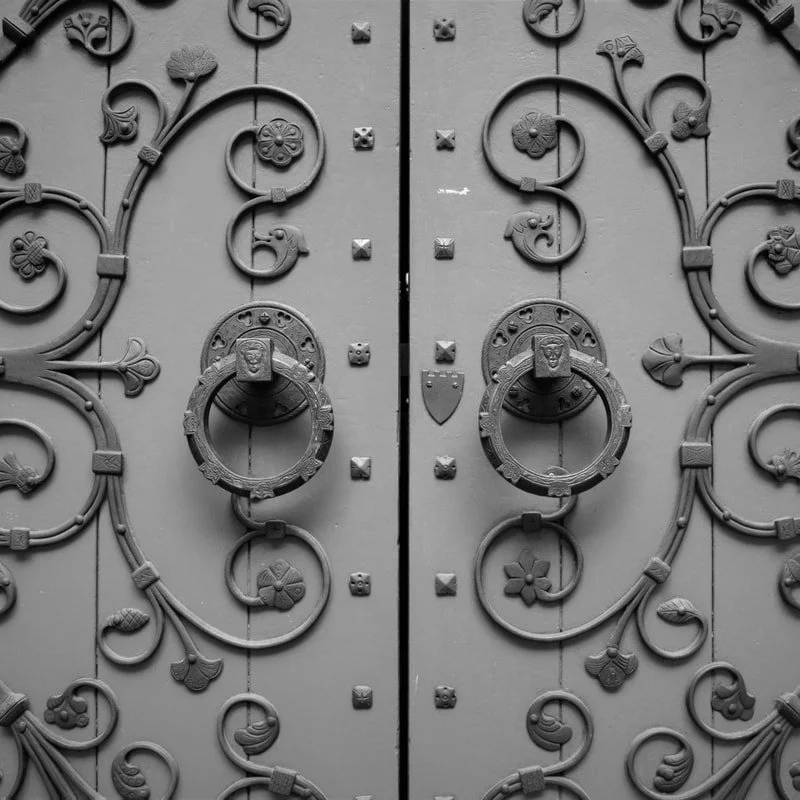 Black and white metal double doors with ornate floral and vine patterns, circular door knockers with faces, and small shield shields.