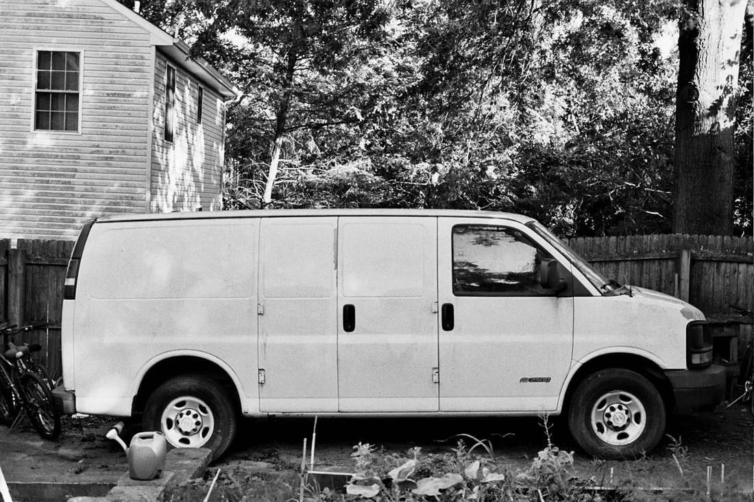 Black and white photo of a white cargo van parked in a backyard with a wooden fence, trees, plants, bicycle, and house in the background.