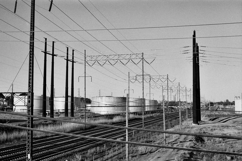 Black and white photo of multiple railway tracks with overhead electrical wires and poles, in an industrial area.