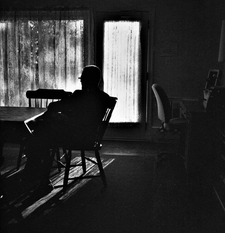 Silhouette of a person sitting at a dining table in a room with window curtains and a sliding glass door, illuminated by natural light from outside.