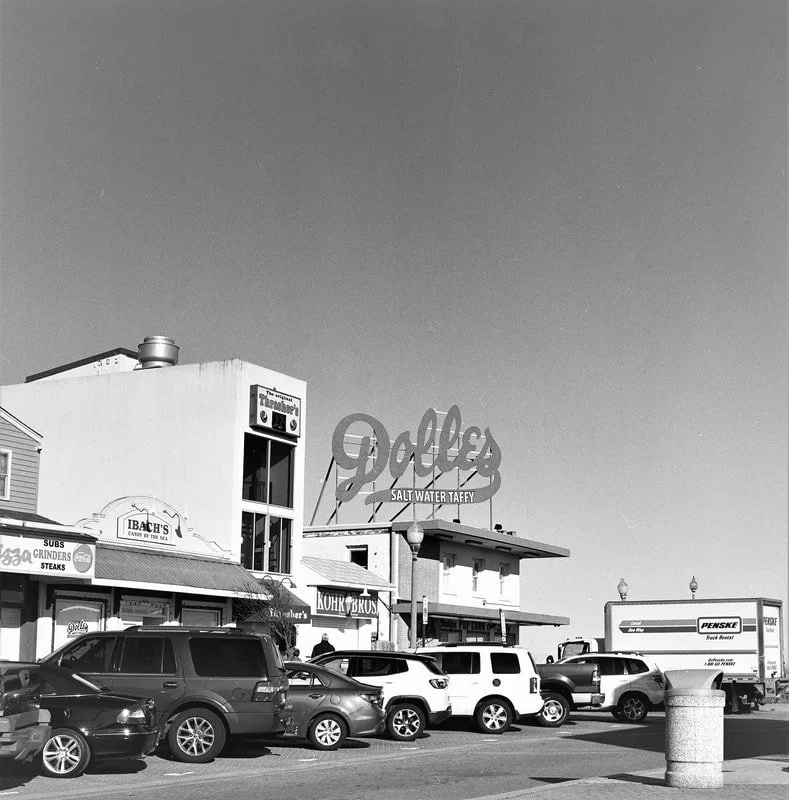 Street scene with parked cars and storefronts, including a diner with a large sign that reads "Dale's Salt Water Taffy" and other businesses in a coastal town.