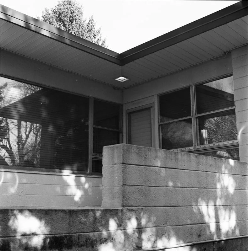 Black and white photo of the corner of a house with large windows, a flat roof, and a concrete block wall.
