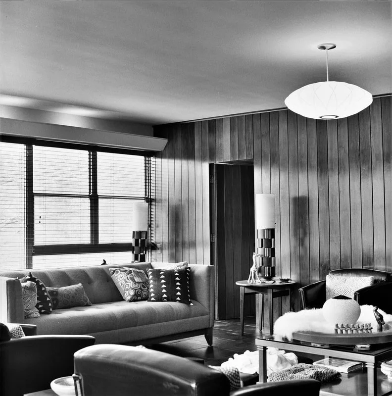 A mid-century modern living room with a large sofa, patterned pillows, a wooden side table with decor, and a coffee table with decorative objects. Wood-paneled walls, large windows with blinds, and a ceiling light fixture are also visible.