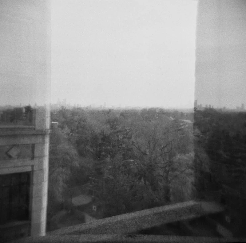 Black and white photo of a cityscape viewed from a window, with trees, buildings, and cloudy sky outside.