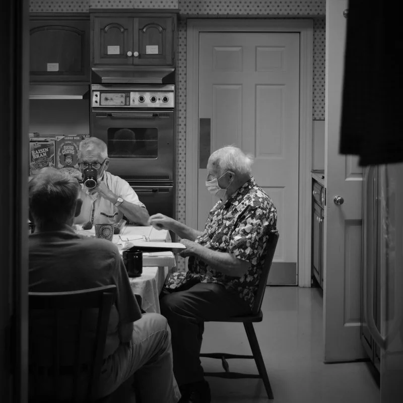 Three elderly men seated at a dining table in a kitchen conversing, with one of them wearing a floral shirt and a face mask, and another drinking from a mug, in black and white.