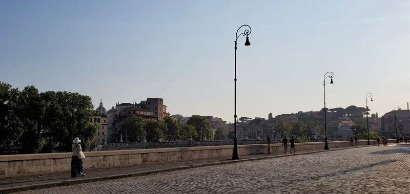 A riverside promenade with cobblestone pavement, tall street lamps, and a woman walking while wearing a white hat. Buildings and trees are visible in the background under a clear sky.