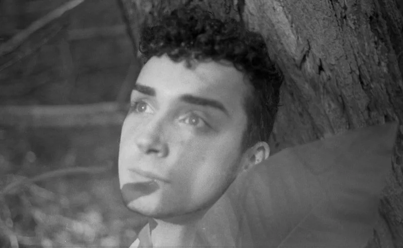 A young person with short curly hair lying next to a tree, looking upward with a thoughtful expression.