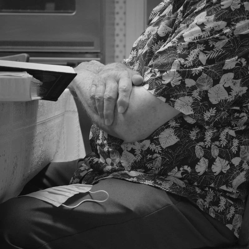 A person sitting with a floral shirt, holding their arm, with a face mask hanging from their lap.