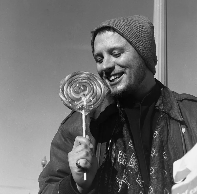 A smiling young man in a beanie holding a large round lollipop.