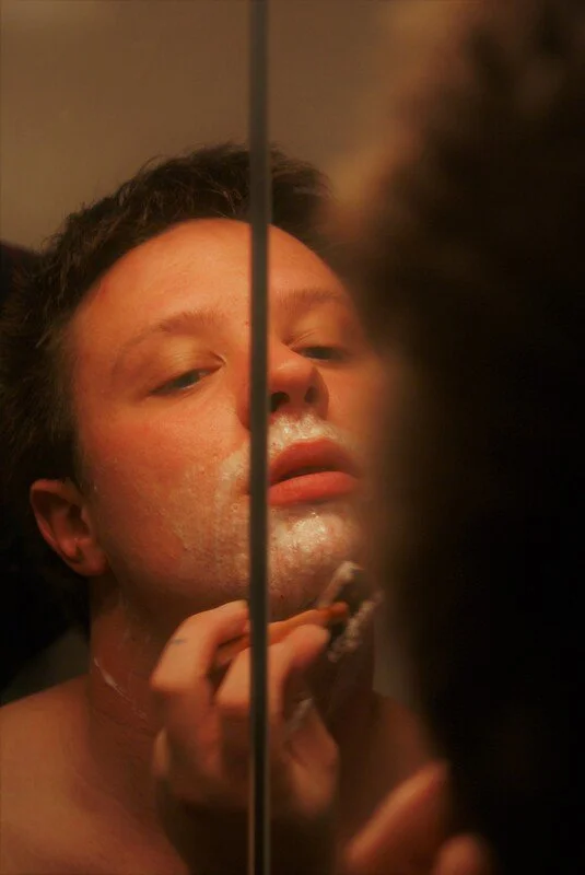 A young man shaving in front of a mirror, seen from a close-up perspective, with a reflection showing his face and hand holding a razor, partially obscured by a blurry dark object in the foreground.