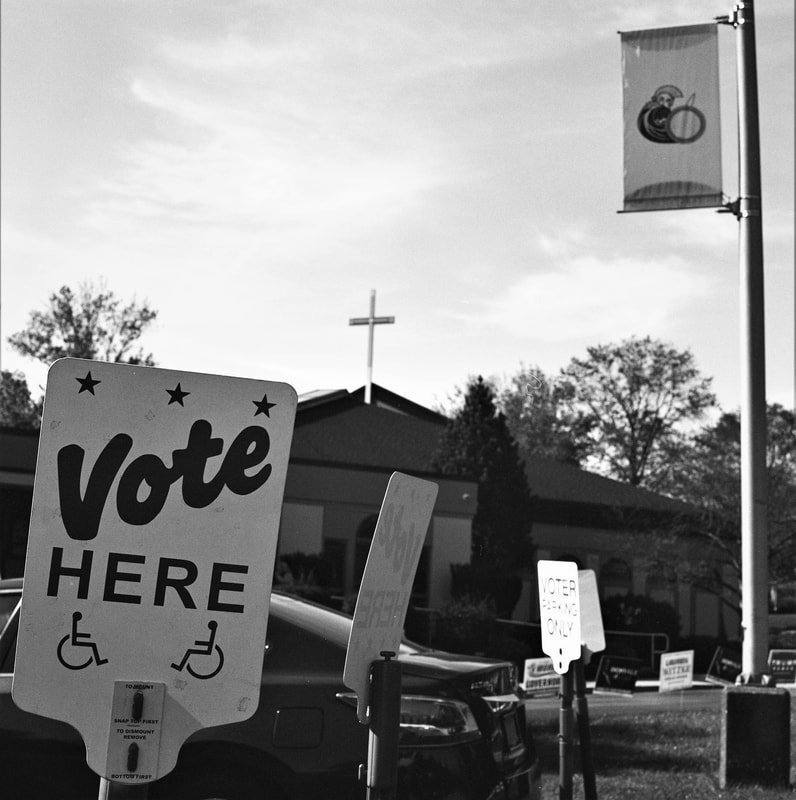 A black and white photo of a voting location with a sign that says 'Vote Here'. In the background, there are small signs and a church with a cross on its roof, trees, cars parked outside, and a flagpole.
