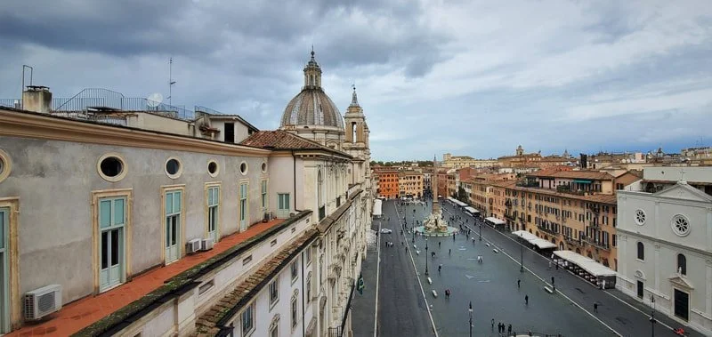 A city street view from above, showing historic buildings, a large domed church, and a fountain in the distance under a cloudy sky.