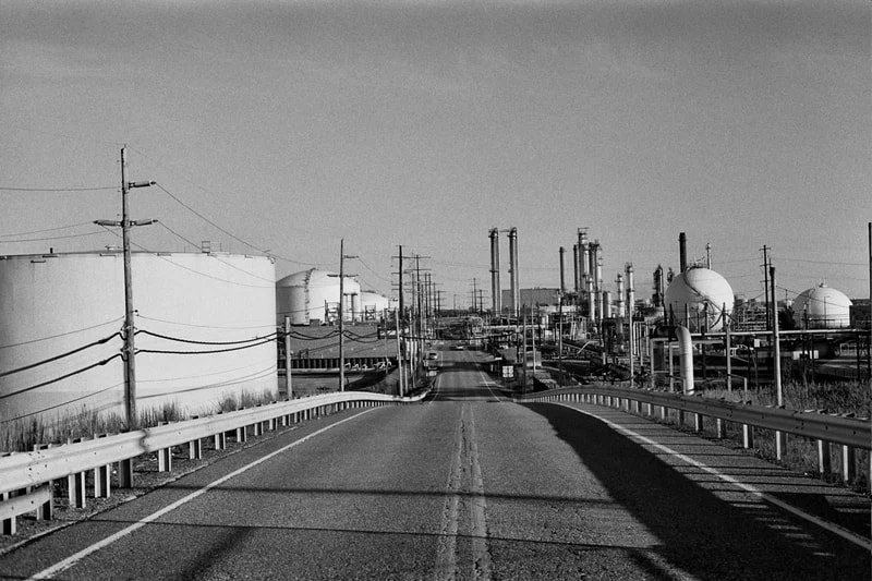 A black and white image of an industrial area with storage tanks and pipes, viewed from a road.