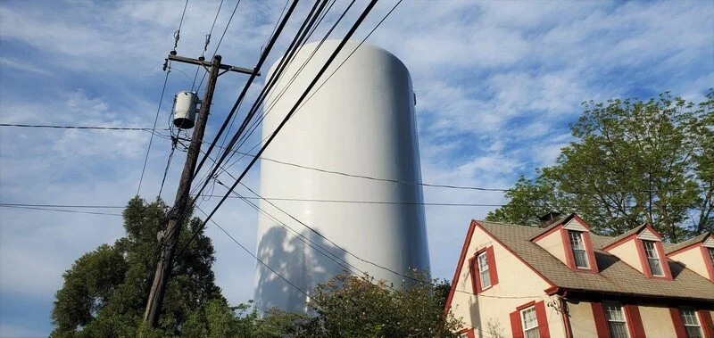 A tall, white silo next to a house with red trim and multiple dormer windows, with trees and a utility pole with wires in the foreground under a partly cloudy sky.