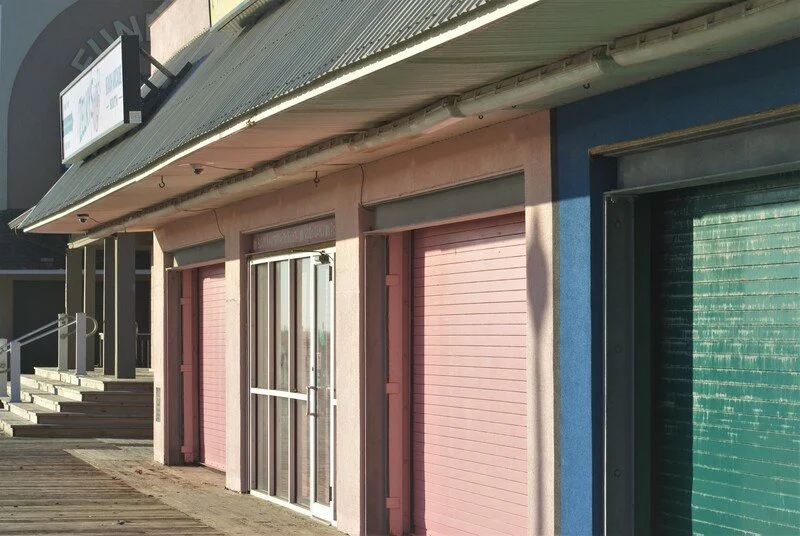 Exterior of a commercial building with closed pink and green roll-up doors, a glass entrance door, and a corrugated metal roof.