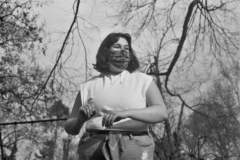 A woman with shoulder-length dark hair and glasses, wearing a face mask and a sleeveless white top, standing outdoors among leafless trees, with a serious expression.