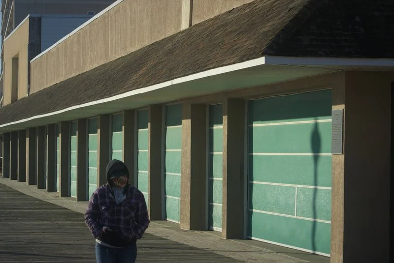A person wearing a mask and hoodie walking along a building with teal-colored garage doors.