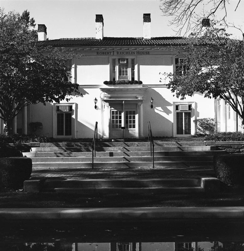 Black and white photo of the Robert J. Reichlin House, a two-story building with a pitched roof and three chimneys. The house has a central entrance with steps, double doors, and decorative outdoor lighting. There are symmetrical windows, trees, and 