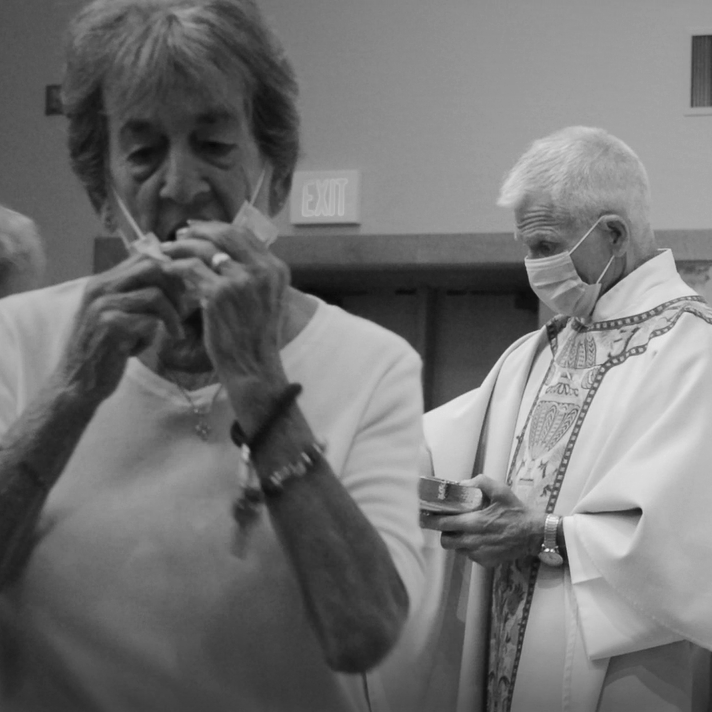 Two elderly women wearing masks, one is lighting a cigarette, and the other is holding a small container, in an indoor setting.