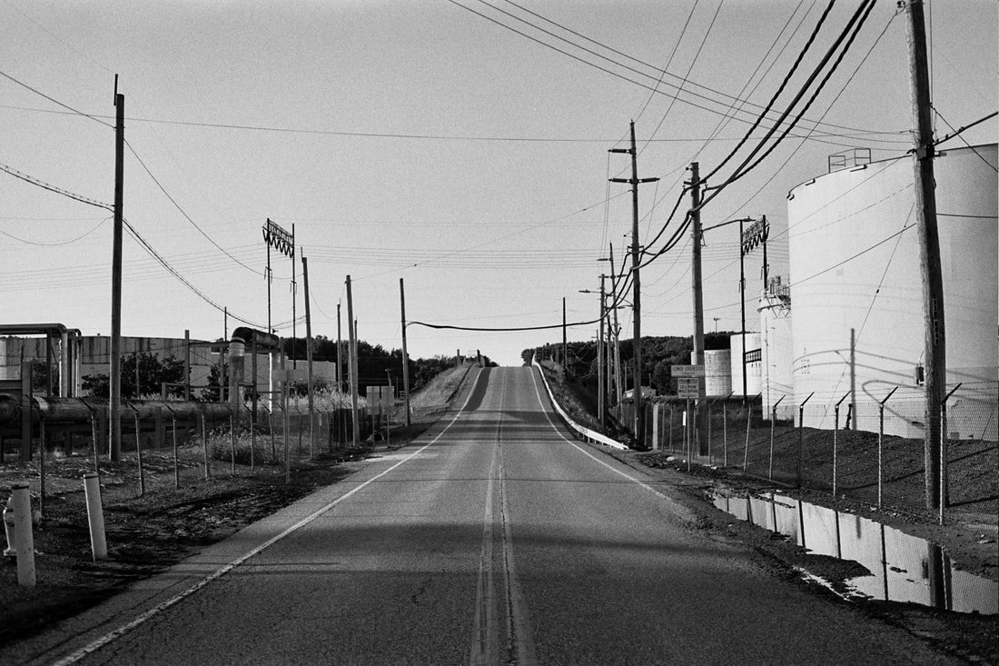 A black-and-white photo of a rural road with power lines on both sides, fencing, and industrial buildings, heading uphill into the distance.