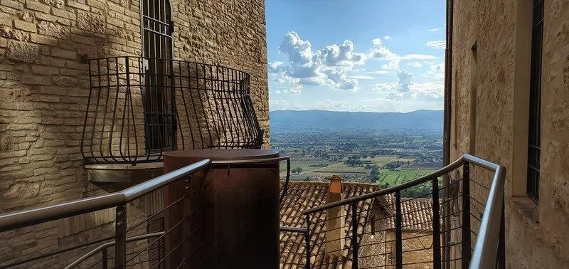 View from a stone balcony overlooking a rural landscape with fields, mountains, and partly cloudy skies.