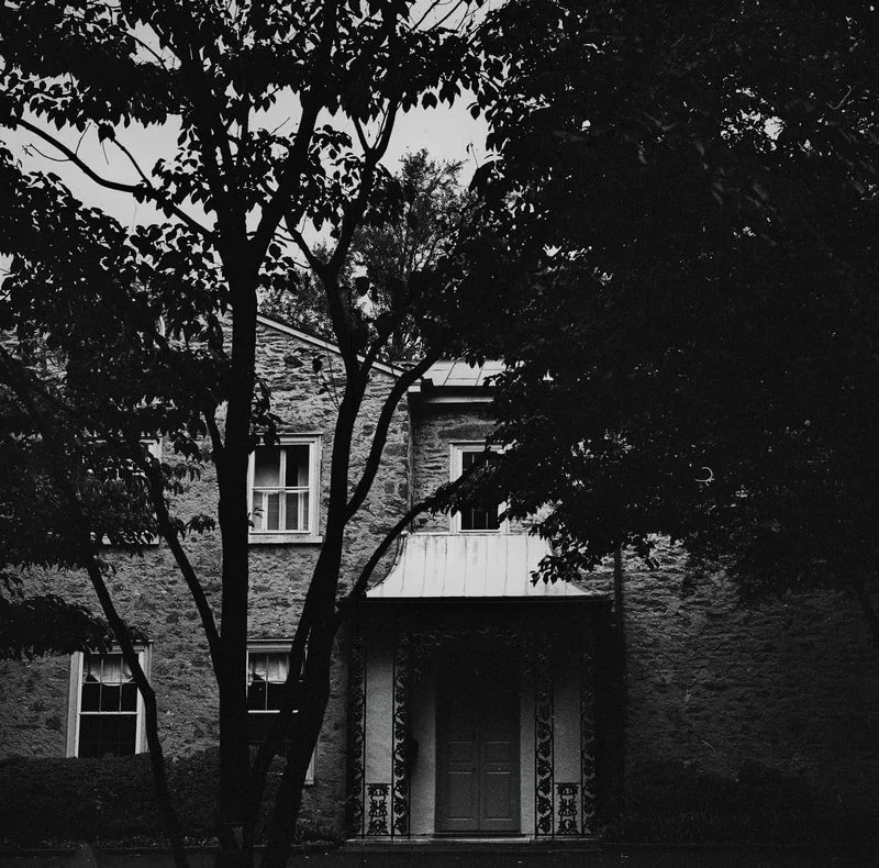 A black and white photo of a house with trees in the foreground, partially obscuring the view of the building.