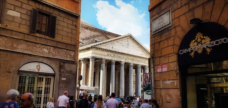 View of the Pantheon in Rome, with people walking and standing in front. Surrounding buildings and a blue sky with some clouds.
