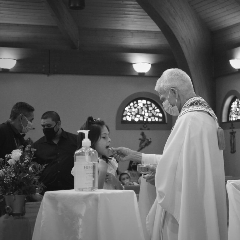 Child receiving communion from priest during a church service with several masked attendees in the background.