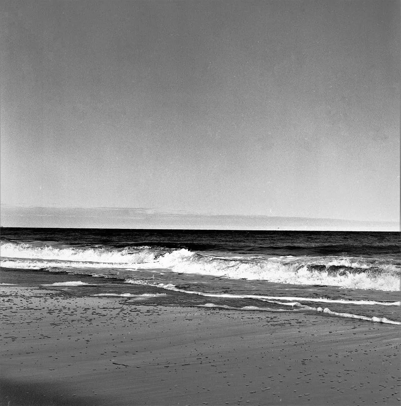 Black and white photo of a calm beach with waves gently crashing onto the sandy shore and a clear sky.